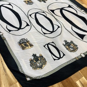 May include: A square, pleated scarf with a white background featuring large black letter "C" designs within oval frames. The scarf also has gold crests and a black border with a gray trim.