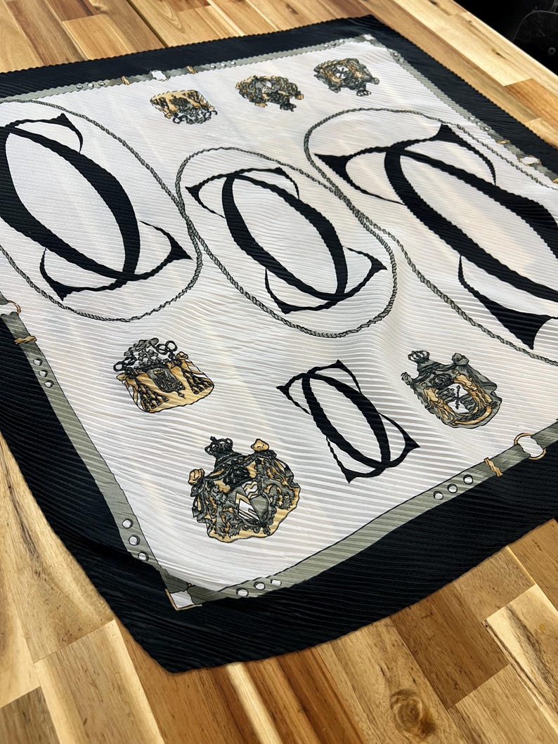 May include: A square, pleated scarf with a white background featuring large black letter "C" designs within oval frames. The scarf also has gold crests and a black border with a gray trim.
