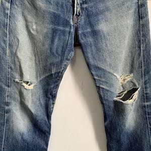 Peut inclure: Jean en denim bleu d&eacute;lav&eacute; avec des d&eacute;tails us&eacute;s, dont des d&eacute;chirures aux genoux. Le jean pr&eacute;sente des signes d'usure, avec des taches plus claires et des coutures visibles.