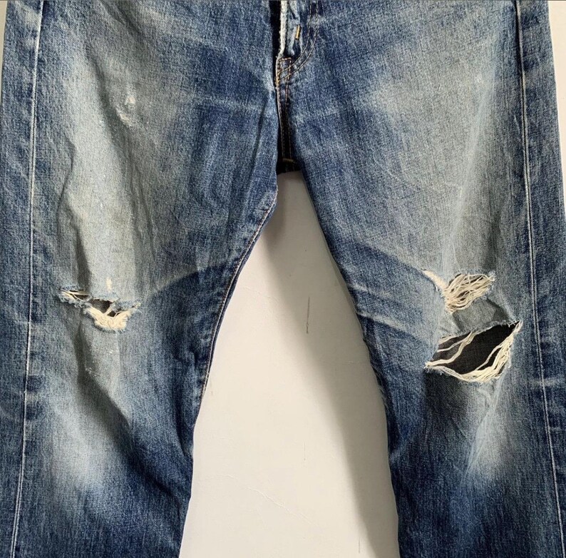 Peut inclure: Jean en denim bleu d&eacute;lav&eacute; avec des d&eacute;tails us&eacute;s, dont des d&eacute;chirures aux genoux. Le jean pr&eacute;sente des signes d'usure, avec des taches plus claires et des coutures visibles.