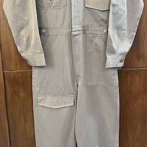May include: A light tan racing jumpsuit with multiple pockets and patches. The jumpsuit features a stand-up collar, long sleeves, and a button-up front. Patches include "SKYLINE RACING TEAM GT-R" and a triangular logo. The garment has a few paint splatters.