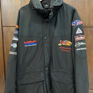 May include: Black Honda Racing jacket with multiple sponsor patches. Features the Honda Racing logo and other racing-related graphics. Includes a hood and multiple pockets. The jacket is hanging on a wooden hanger.