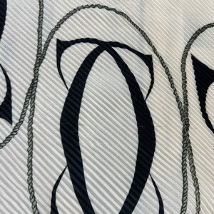 May include: Close-up of a white fabric with a pleated texture. The fabric features a repeating pattern of black abstract shapes and gray rope-like designs. The background has thin, vertical gray stripes.