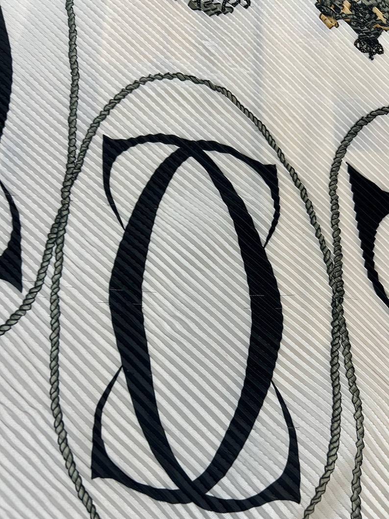 May include: Close-up of a white fabric with a pleated texture. The fabric features a repeating pattern of black abstract shapes and gray rope-like designs. The background has thin, vertical gray stripes.