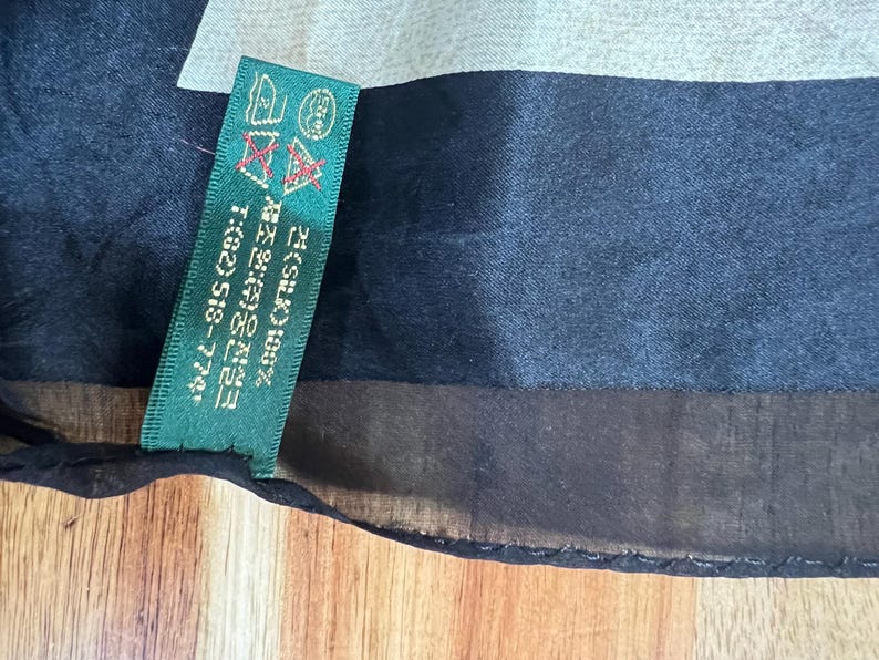 May include: A black silk scarf with a green tag. The tag has text in Korean and English, including "SILK 100%" and a phone number. The scarf has a dark blue and light grey pattern.