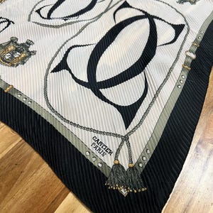 May include: A pleated silk scarf with a repeating pattern of black "C" shapes and rope detailing on a white background. The scarf has a black border with a gray and gold accent. The text "CARTIER PARIS" is visible.