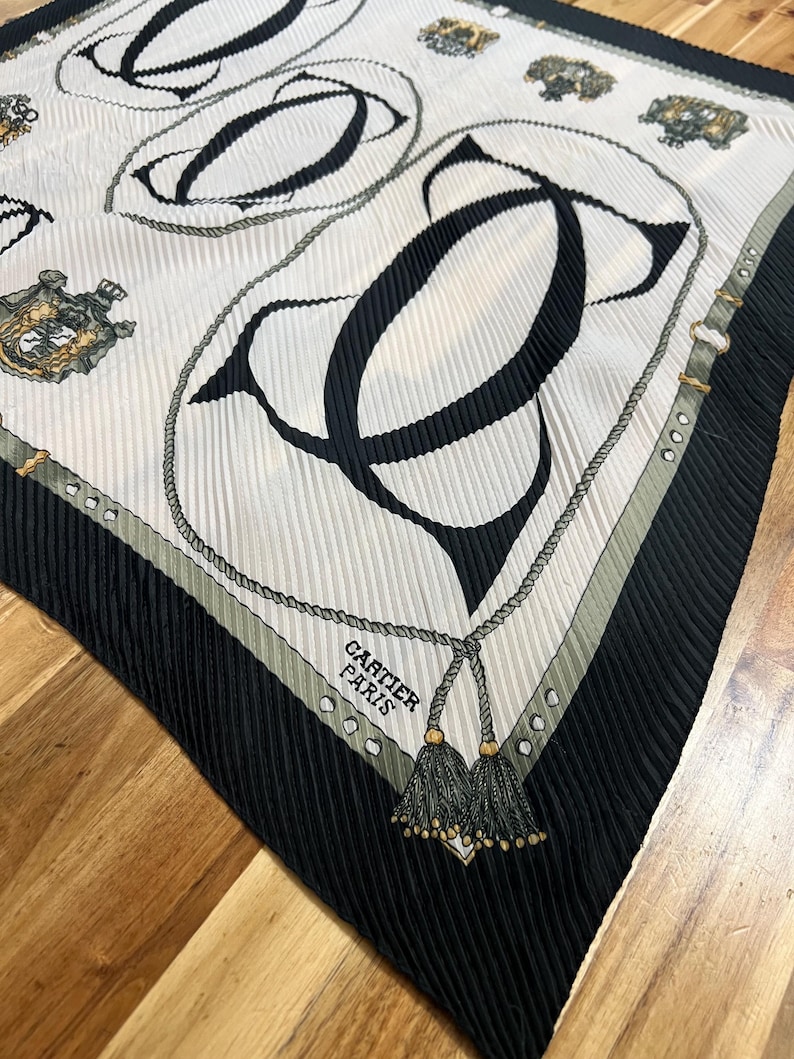 May include: A pleated silk scarf with a repeating pattern of black "C" shapes and rope detailing on a white background. The scarf has a black border with a gray and gold accent. The text "CARTIER PARIS" is visible.