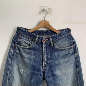 Peut inclure: Un jean en denim bleu suspendu &agrave; un cintre en bois. Le jean pr&eacute;sente une conception classique &agrave; cinq poches, une fermeture boutonn&eacute;e et un d&eacute;lavage d&eacute;color&eacute;. Le denim montre des signes d'usure, avec des taches plus claires et une d&eacute;tresse subtile.