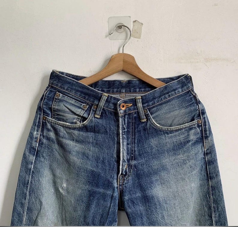 Peut inclure: Un jean en denim bleu suspendu &agrave; un cintre en bois. Le jean pr&eacute;sente une conception classique &agrave; cinq poches, une fermeture boutonn&eacute;e et un d&eacute;lavage d&eacute;color&eacute;. Le denim montre des signes d'usure, avec des taches plus claires et une d&eacute;tresse subtile.