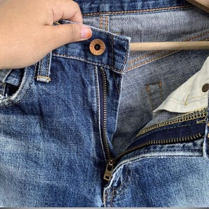 Peut inclure: Gros plan sur un jean en denim bleu avec une fermeture &eacute;clair partiellement ouverte. Le jean a une fermeture boutonn&eacute;e avec le nom de la marque visible. Le denim montre des signes d'usure, avec un aspect d&eacute;lav&eacute;. La fermeture &eacute;clair est dor&eacute;e.