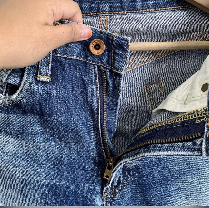 Peut inclure: Gros plan sur un jean en denim bleu avec une fermeture &eacute;clair partiellement ouverte. Le jean a une fermeture boutonn&eacute;e avec le nom de la marque visible. Le denim montre des signes d'usure, avec un aspect d&eacute;lav&eacute;. La fermeture &eacute;clair est dor&eacute;e.