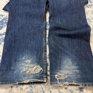Może przedstawiać: Ciemnoniebieskie dżinsy z denimu z przetarciami, w tym rozdarciami i wyblakłymi obszarami. Dżinsy mają klasyczny design z pięcioma kieszeniami i brązową sk&oacute;rzaną naszywką z tyłu. Dżinsy są złożone, pokazując prz&oacute;d i tył.