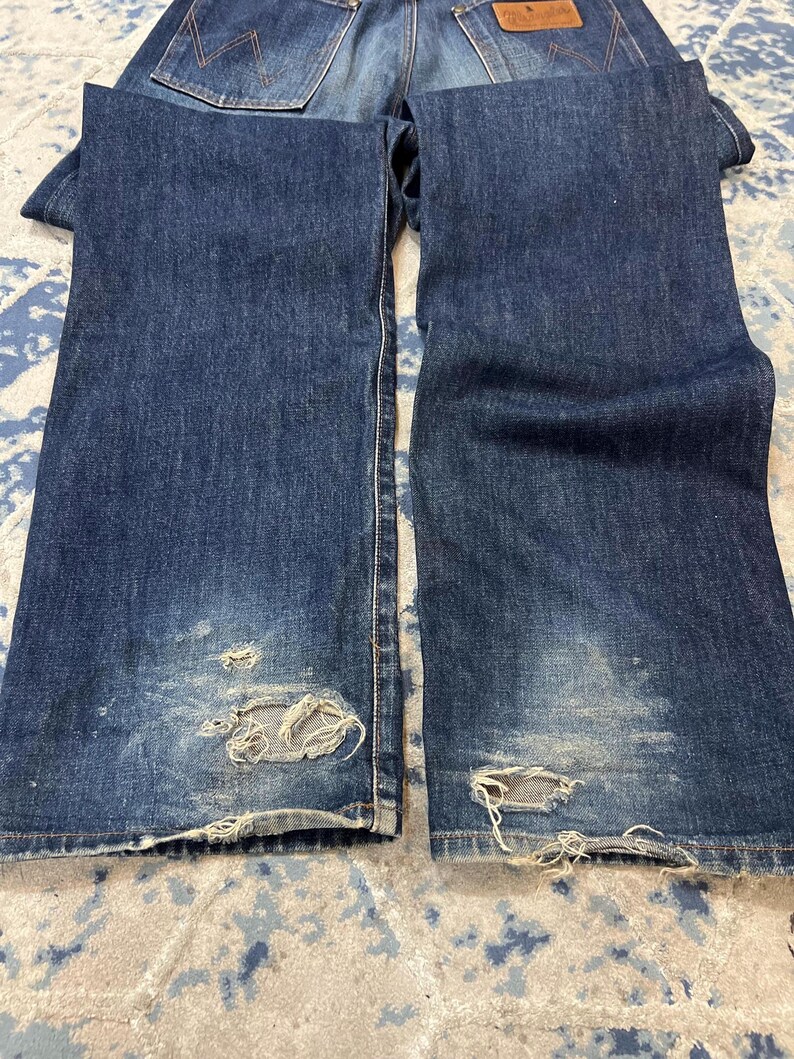 Może przedstawiać: Ciemnoniebieskie dżinsy z denimu z przetarciami, w tym rozdarciami i wyblakłymi obszarami. Dżinsy mają klasyczny design z pięcioma kieszeniami i brązową sk&oacute;rzaną naszywką z tyłu. Dżinsy są złożone, pokazując prz&oacute;d i tył.