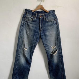 Peut inclure: Jean bleu d&eacute;lav&eacute; avec des d&eacute;tails us&eacute;s, dont des d&eacute;chirures aux genoux. Le jean est suspendu &agrave; un cintre en bois contre un mur blanc. Le jean a une fermeture &agrave; bouton et une conception classique &agrave; cinq poches.