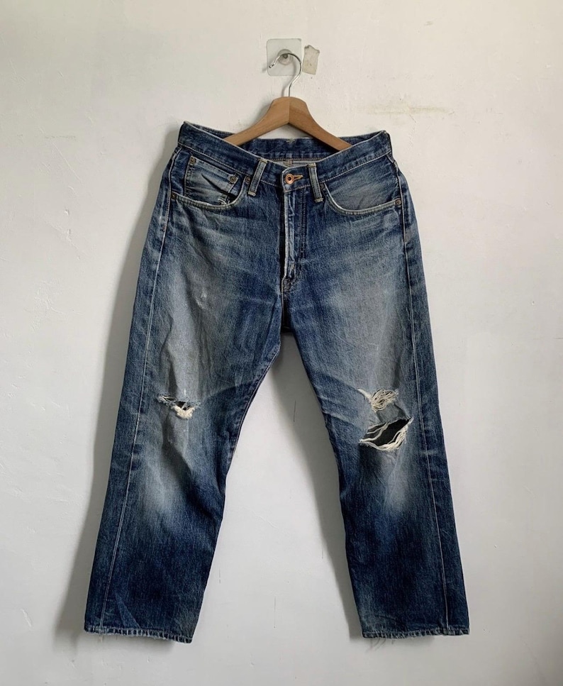 Peut inclure: Jean bleu d&eacute;lav&eacute; avec des d&eacute;tails us&eacute;s, dont des d&eacute;chirures aux genoux. Le jean est suspendu &agrave; un cintre en bois contre un mur blanc. Le jean a une fermeture &agrave; bouton et une conception classique &agrave; cinq poches.