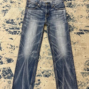Könnte beinhalten: Eine blaue Jeans aus Denim mit einem verwaschenen Whisker-Muster auf den Beinen. Die Hose verfügt über einen klassischen Knopfverschluss, fünf Taschen und einen geradlinigen Schnitt mit sichtbaren Nähten entlang der Beine und am Bund.