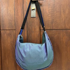 K&ouml;nnte beinhalten: Eine gro&szlig;e, schillernde Umh&auml;ngetasche in T&uuml;rkis-, Lila- und Gr&uuml;nt&ouml;nen h&auml;ngt an einem T&uuml;rgriff. Die Tasche hat eine Halbmondform mit einem schwarzen und blauen Riemen. Das Material der Tasche hat ein strukturiertes, zerknittertes Aussehen.