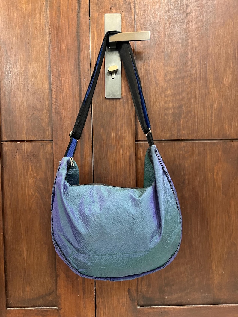 K&ouml;nnte beinhalten: Eine gro&szlig;e, schillernde Umh&auml;ngetasche in T&uuml;rkis-, Lila- und Gr&uuml;nt&ouml;nen h&auml;ngt an einem T&uuml;rgriff. Die Tasche hat eine Halbmondform mit einem schwarzen und blauen Riemen. Das Material der Tasche hat ein strukturiertes, zerknittertes Aussehen.