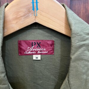 May include: An olive green shirt with a textured fabric finish. A rectangular red label is stitched at the collar, featuring the text PX Avirex Authentic Garment in white and black. A small white tag below the label displays the size M.