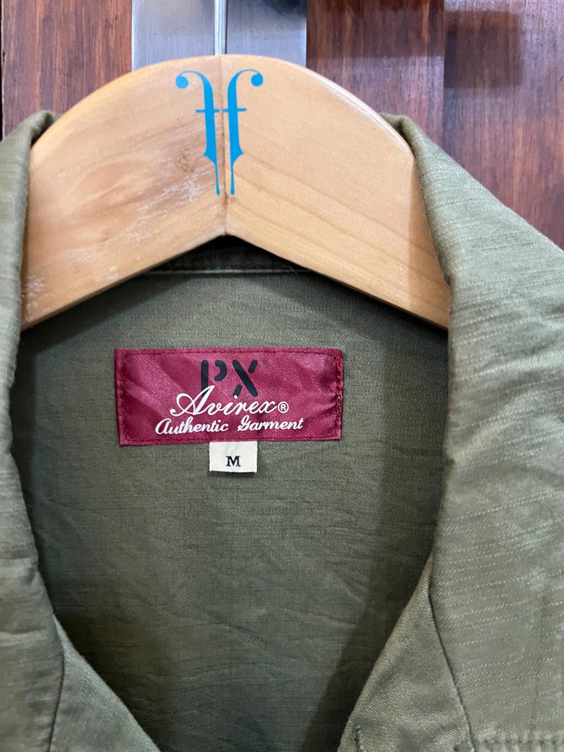 May include: An olive green shirt with a textured fabric finish. A rectangular red label is stitched at the collar, featuring the text PX Avirex Authentic Garment in white and black. A small white tag below the label displays the size M.