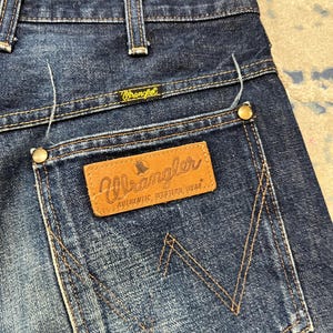 Może przedstawiać: Zbliżenie na parę niebieskich dżins&oacute;w Wrangler. Dżinsy mają klasyczny pięciokieszeniowy design z brązową sk&oacute;rzaną naszywką na tylnej kieszeni. Logo Wrangler jest widoczne na naszywce i nad tylną kieszenią.