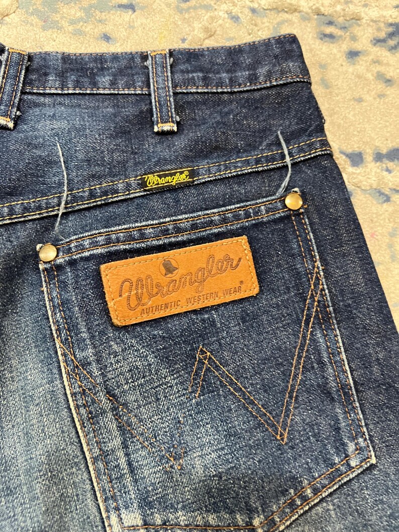 Może przedstawiać: Zbliżenie na parę niebieskich dżins&oacute;w Wrangler. Dżinsy mają klasyczny pięciokieszeniowy design z brązową sk&oacute;rzaną naszywką na tylnej kieszeni. Logo Wrangler jest widoczne na naszywce i nad tylną kieszenią.