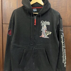 Pu&ograve; includere: Felpa nera con zip, cappuccio e tasche anteriori. Presenta una grafica di geisha e la scritta "Defiance" sul davanti. La manica destra ha testo e un disegno.