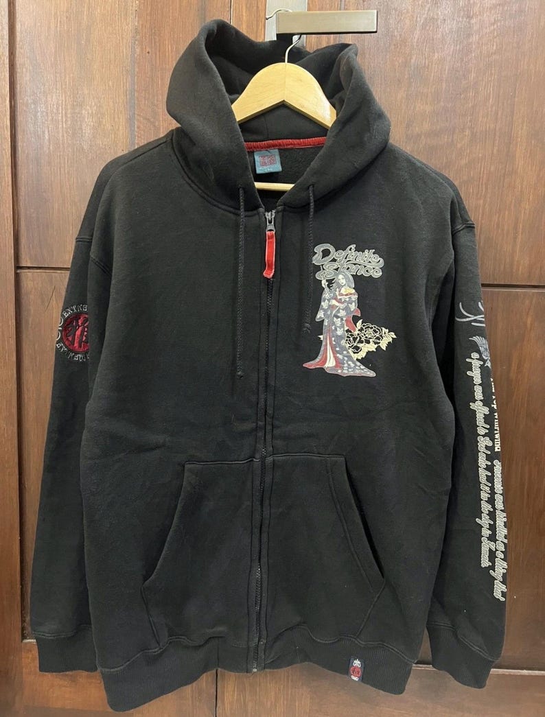 Pu&ograve; includere: Felpa nera con zip, cappuccio e tasche anteriori. Presenta una grafica di geisha e la scritta "Defiance" sul davanti. La manica destra ha testo e un disegno.