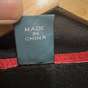 Pu&ograve; includere: Primo piano di un'etichetta turchese con la scritta bianca "MADE IN CHINA". L'etichetta &egrave; attaccata a un capo nero con una cucitura rossa. L'immagine mostra i dettagli dell'etichetta e del tessuto.