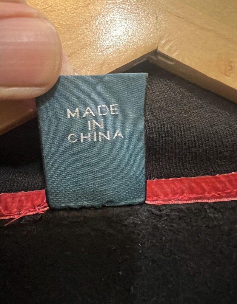 Pu&ograve; includere: Primo piano di un'etichetta turchese con la scritta bianca "MADE IN CHINA". L'etichetta &egrave; attaccata a un capo nero con una cucitura rossa. L'immagine mostra i dettagli dell'etichetta e del tessuto.