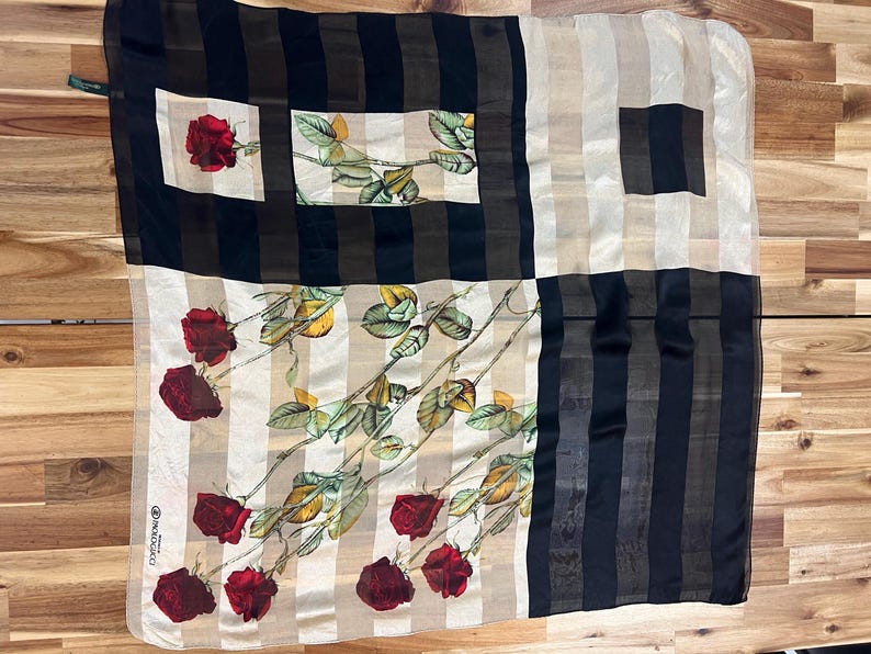 May include: A square silk scarf with a floral rose pattern. The scarf features a design of red roses and green leaves on a cream and beige striped background, with black and brown vertical stripes.