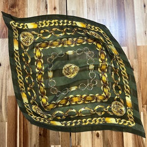 May include: A square, olive green scarf with a gold chain and medallion pattern. The scarf has a pleated texture and a dark green border. The gold chains and medallions create a luxurious and elegant design.