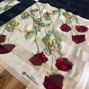 May include: A square silk scarf with a floral rose design. The scarf features a cream-colored background with vertical stripes, black panels, and red roses with green leaves. The text "DESIGNED BY PAOLOGUCCI" is printed on the scarf.