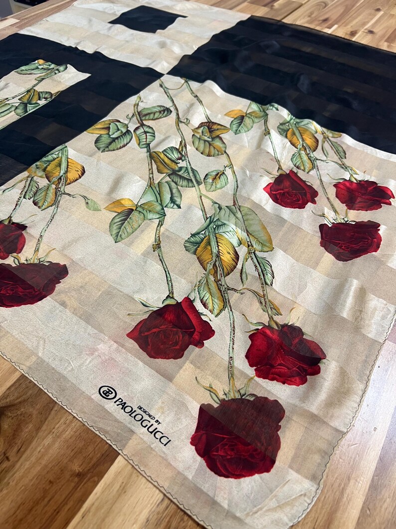 May include: A square silk scarf with a floral rose design. The scarf features a cream-colored background with vertical stripes, black panels, and red roses with green leaves. The text "DESIGNED BY PAOLOGUCCI" is printed on the scarf.