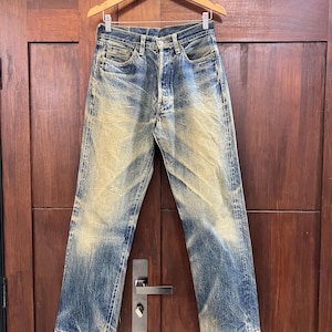 May include: A pair of blue denim jeans featuring a heavy distressed wash with faded yellow and white highlights, a classic five-pocket design, a button fly closure, and frayed hems at the bottom of the straight-cut legs, hanging on a wooden hanger.