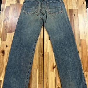 May include: Faded blue denim jeans with a classic five-pocket design. The jeans have a straight leg fit and a worn appearance, with a leather patch on the back waistband. The denim shows signs of wear and tear, adding to the vintage aesthetic.