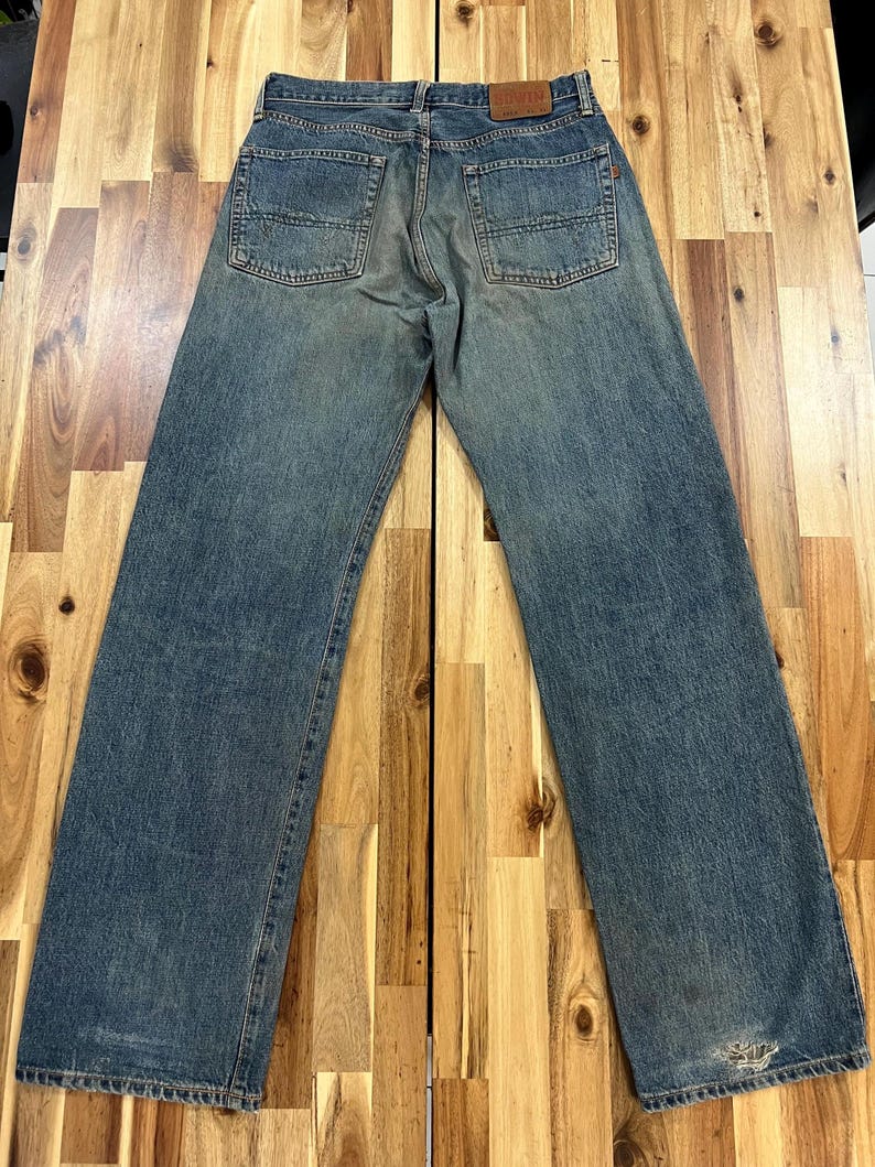 May include: Faded blue denim jeans with a classic five-pocket design. The jeans have a straight leg fit and a worn appearance, with a leather patch on the back waistband. The denim shows signs of wear and tear, adding to the vintage aesthetic.