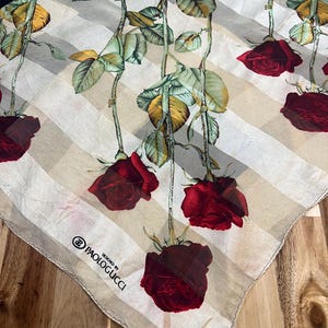 May include: A square silk scarf featuring a design of red roses with green leaves and stems against a cream and white striped background. The scarf has a black border and the text "PAOLOGUCCI DESIGNED BY" is printed on the corner.