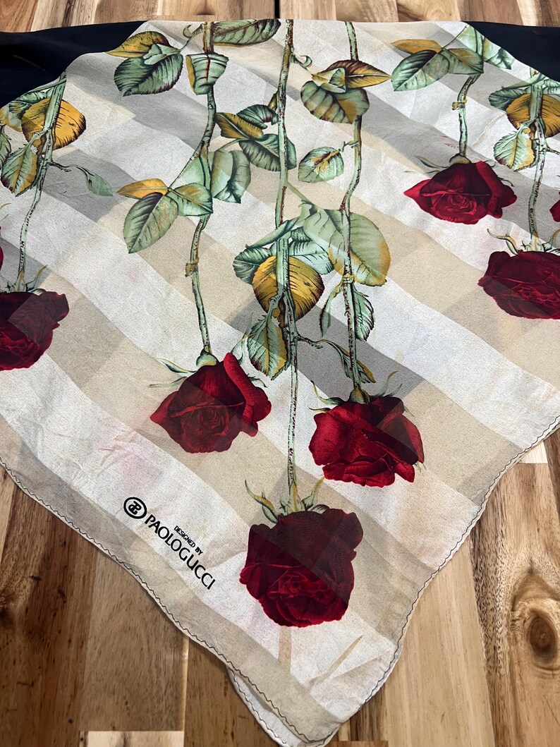 May include: A square silk scarf featuring a design of red roses with green leaves and stems against a cream and white striped background. The scarf has a black border and the text "PAOLOGUCCI DESIGNED BY" is printed on the corner.