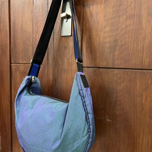 K&ouml;nnte beinhalten: Eine blau-violett schillernde Umh&auml;ngetasche mit schwarzem Riemen und Rei&szlig;verschluss. Die Tasche h&auml;ngt an einer Holzt&uuml;r. Die Tasche hat eine Halbmondform und ein schwarzes Etikett.