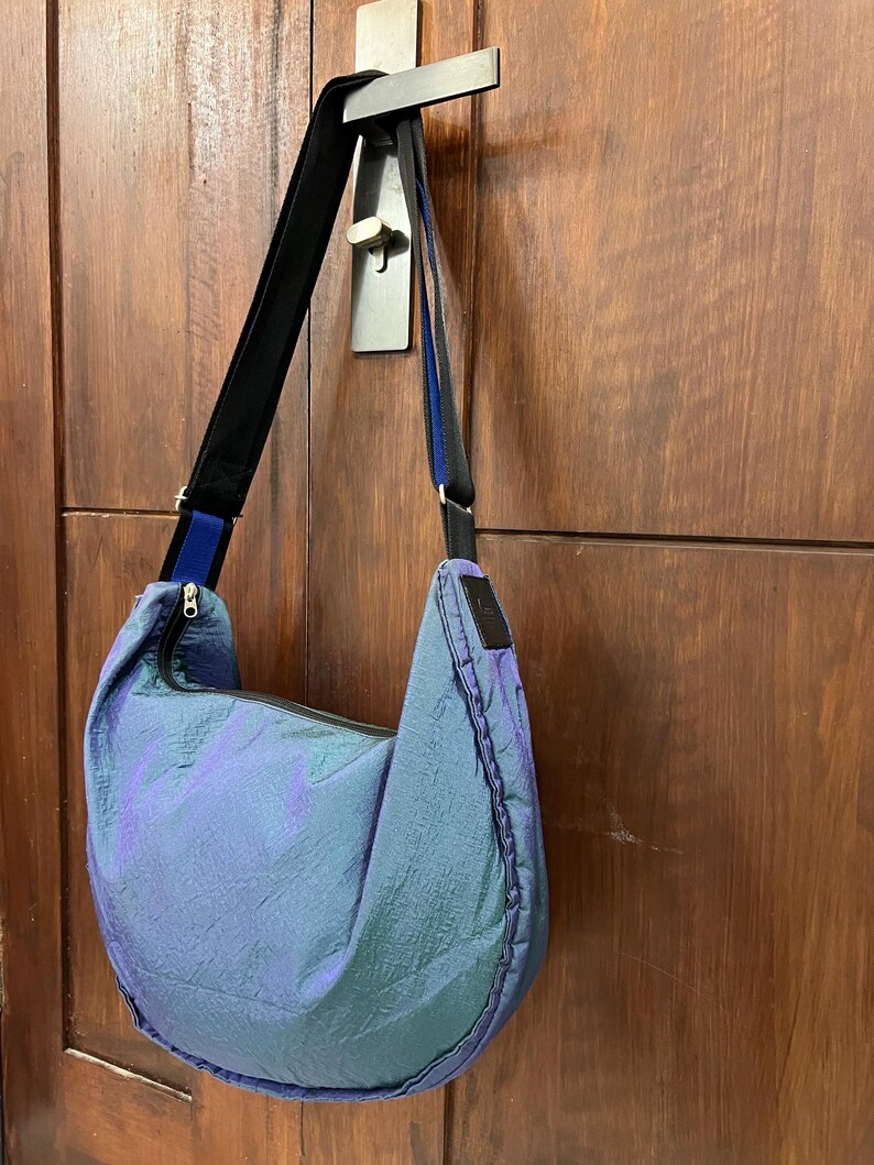 K&ouml;nnte beinhalten: Eine blau-violett schillernde Umh&auml;ngetasche mit schwarzem Riemen und Rei&szlig;verschluss. Die Tasche h&auml;ngt an einer Holzt&uuml;r. Die Tasche hat eine Halbmondform und ein schwarzes Etikett.