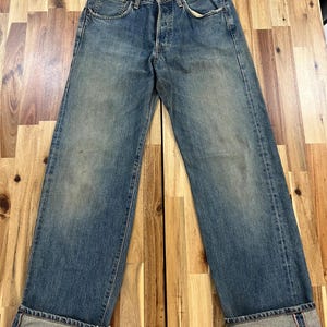 May include: Faded blue denim jeans with a button fly and classic five-pocket design. The jeans have a relaxed fit and are cuffed at the ankles, revealing a red selvedge detail. The denim shows signs of wear and fading.