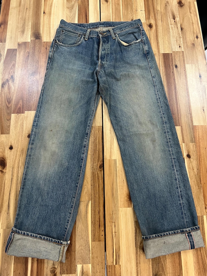 May include: Faded blue denim jeans with a button fly and classic five-pocket design. The jeans have a relaxed fit and are cuffed at the ankles, revealing a red selvedge detail. The denim shows signs of wear and fading.
