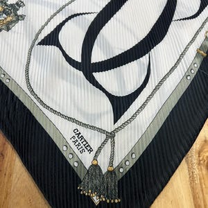 May include: A silk scarf with a pleated texture, featuring a black and white design. The scarf has a large black "C" shape, rope detailing, and the text "CARTIER PARIS". The edges are black with a gray border and gold tassels.