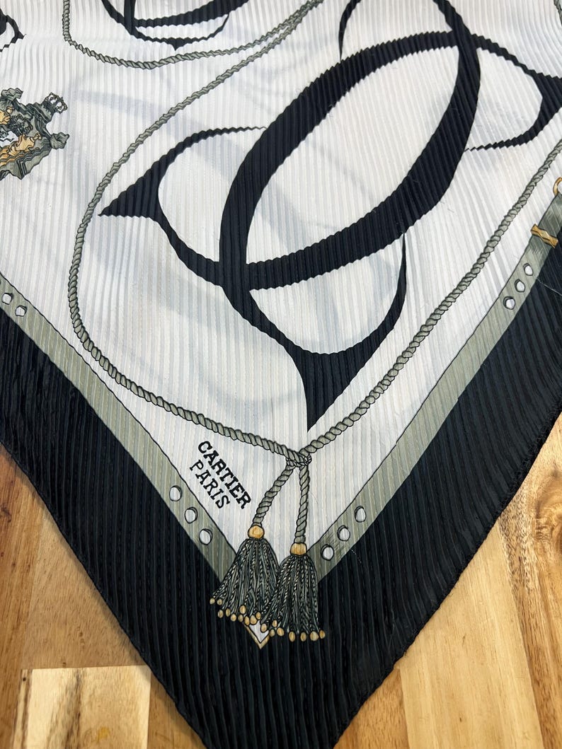May include: A silk scarf with a pleated texture, featuring a black and white design. The scarf has a large black "C" shape, rope detailing, and the text "CARTIER PARIS". The edges are black with a gray border and gold tassels.