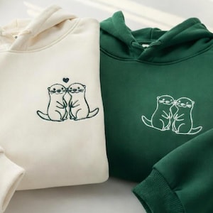 Könnte beinhalten: Zwei Kapuzenpullover, einer in Creme und einer in Dunkelgrün, jeweils mit einer Stickerei von zwei händchenhaltenden Ottern. Der cremefarbene Hoodie zeigt die Otter mit einem kleinen Herz darüber, während der grüne Hoodie die Otter nebeneinander zeigt.