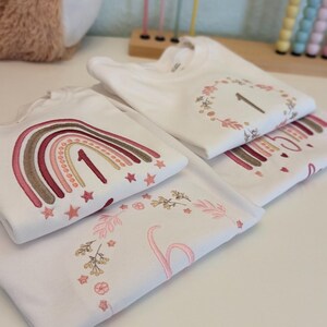 May include: Three white t-shirts with embroidered designs. The first shirt features a rainbow with stars and the number "1". The second shirt has a wreath with leaves and the number "1". The third shirt has a floral design with the number "1".
