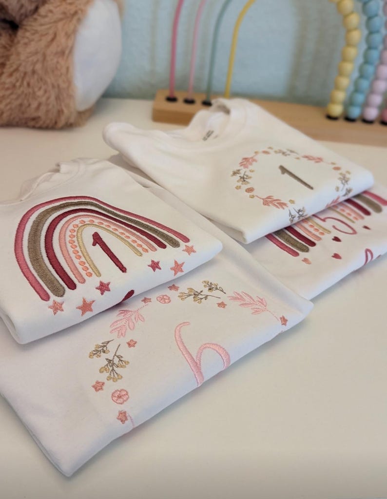 May include: Three white t-shirts with embroidered designs. The first shirt features a rainbow with stars and the number "1". The second shirt has a wreath with leaves and the number "1". The third shirt has a floral design with the number "1".