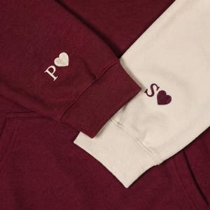 Op de afbeelding: Bordeaux hoodie met crèmekleurige mouwen. De linker manchet heeft de geborduurde letter "P" naast een witte hart. De rechter manchet heeft de geborduurde letter "S" naast een bordeauxrood hart. De hoodie is gemaakt van een zacht materiaal.