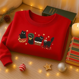 Op de afbeelding: Een rode sweatshirt met een geborduurd ontwerp van vier zwarte katten in feestelijke kleding. De katten worden afgebeeld met een geschenkdoos, krans en kerstmuts. De sweatshirt heeft een ronde hals en lange mouwen.
